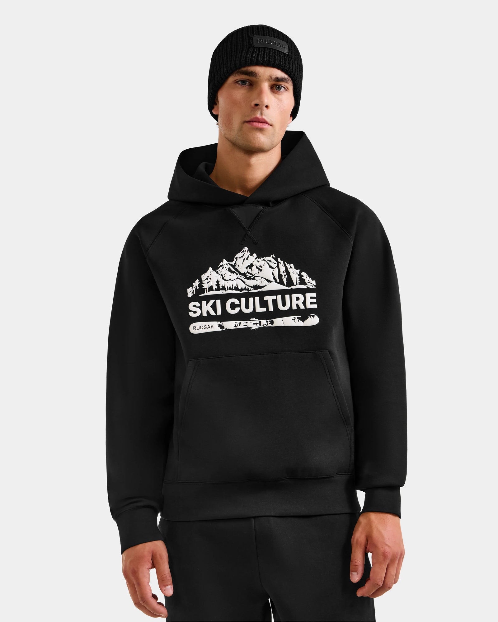 A man in a black beanie and the Rudsak DARWIN - M525502 Black men’s hoodie, featuring a white SKI CULTURE mountain graphic and crafted from premium cotton-blend fleece, stands against a plain light background.
