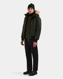 A man wears the Rudsak DEACON - M123513 Dark Olive men's winter coat with a fur-trimmed hood, paired with black pants, boots, and a dark knit beanie against a plain light gray background.