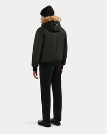 A person stands facing away against a plain light background, wearing the Rudsak DEACON - M123513 Dark Olive bomber-style down puffer, paired with black pants, black shoes, and a black knit beanie.