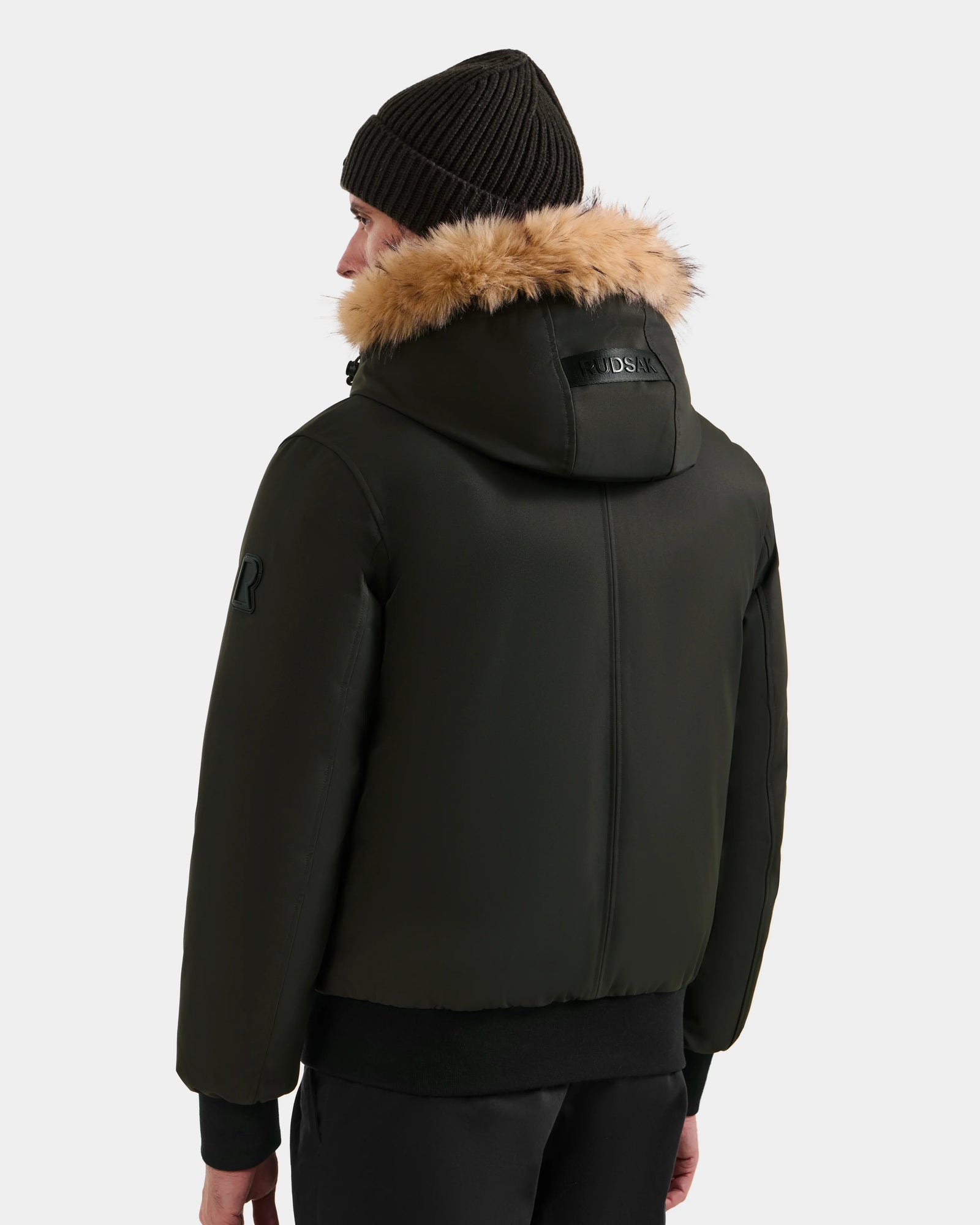 A person wearing the Rudsak DEACON - M123513 men's down puffer coat in dark olive with light brown fur trim, a black knit beanie, and black pants, shown from the back against a plain light background.