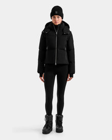 A person stands facing forward, wearing the Rudsak DELILAH M - L125538 Black women’s down puffer with removable sleeves, paired with black leggings, a black beanie, and black boots against a plain light gray background.