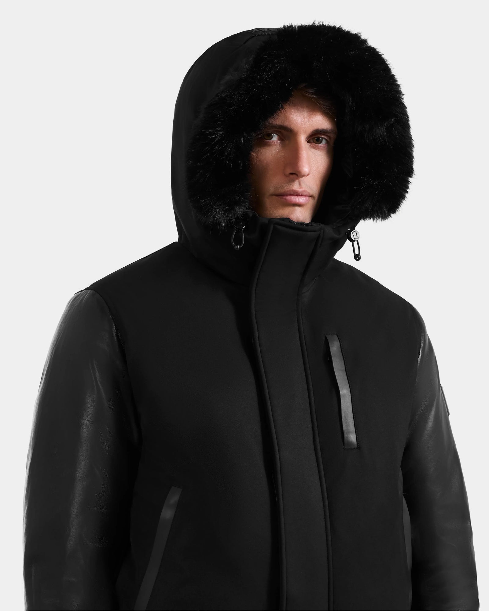 A person wears the Rudsak DERON - M125539 Black men’s down bomber coat with RDS-certified fill, vegan-leather sleeves, a fur-trimmed hood, and front zipper, looking forward with a neutral expression.