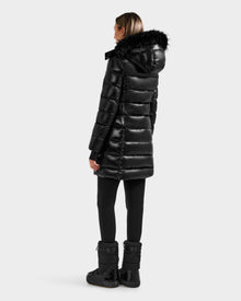 A person stands facing away, wearing the Rudsak ELISA S - L125572 Black women's down puffer with a fur-trimmed hood, paired with black leggings and winter boots against a plain light background.