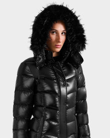 A person wearing the Rudsak ELISA S - L125572 Black puffer jacket with a shiny quilted finish and large black faux fur-trimmed hood, standing against a plain light gray background.