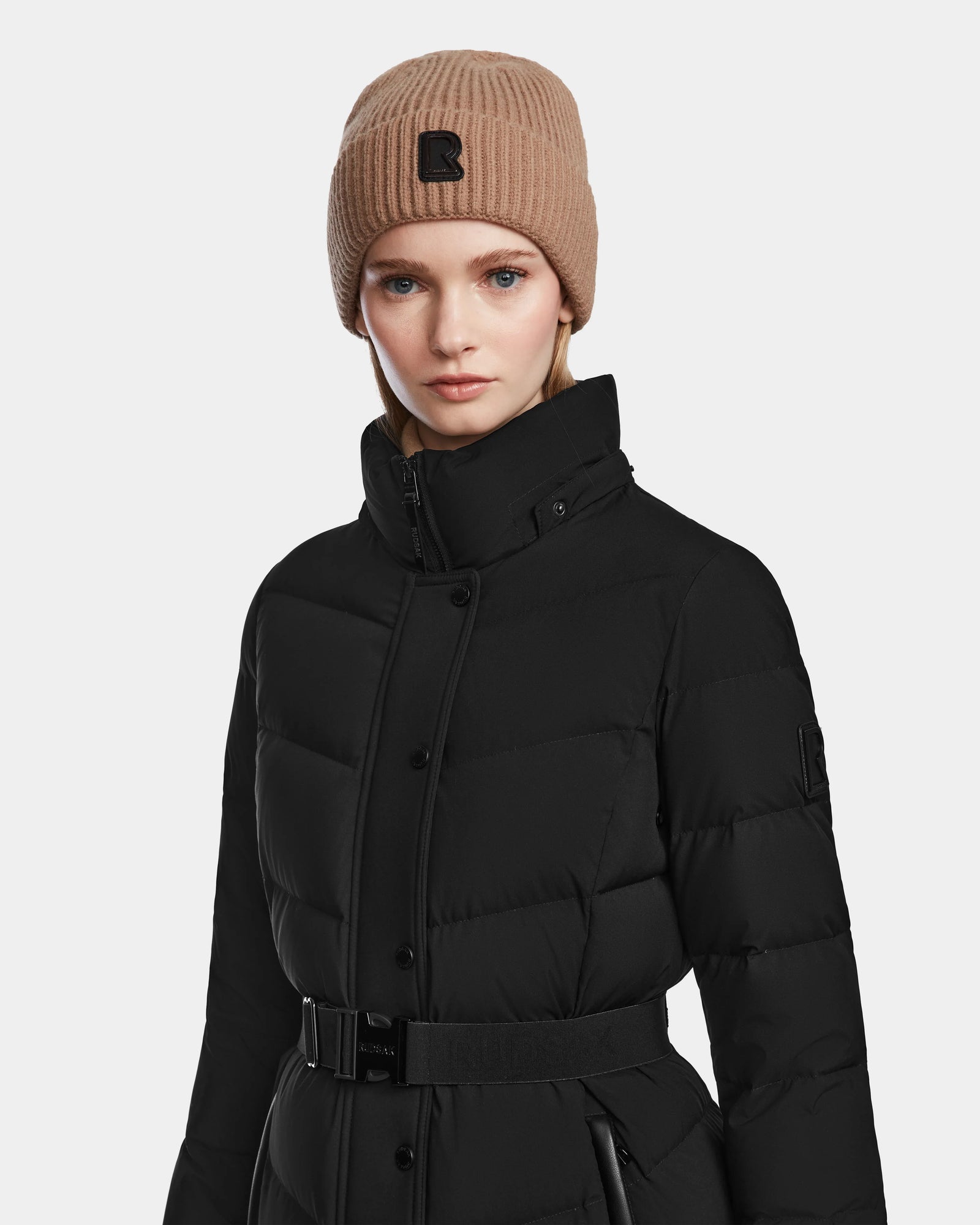 A woman in the Rudsak ELSIE M - L123517 Black/Natural belted maxi down puffer coat and a tan knit beanie with a black patch poses against a plain light background, maintaining a neutral expression.
