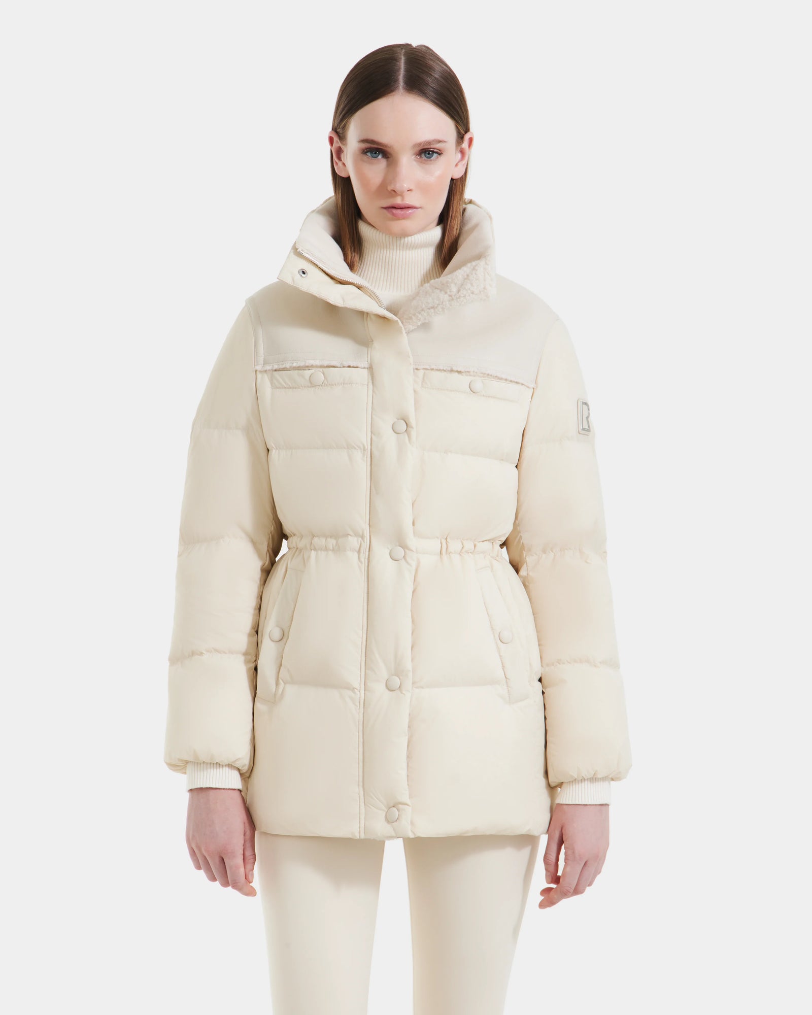 A woman in the Rudsak EMBER M-M - L125524 Vanilla light beige down puffer jacket with a high collar and front buttons, paired with matching beige pants and a turtleneck, stands against a plain white background.