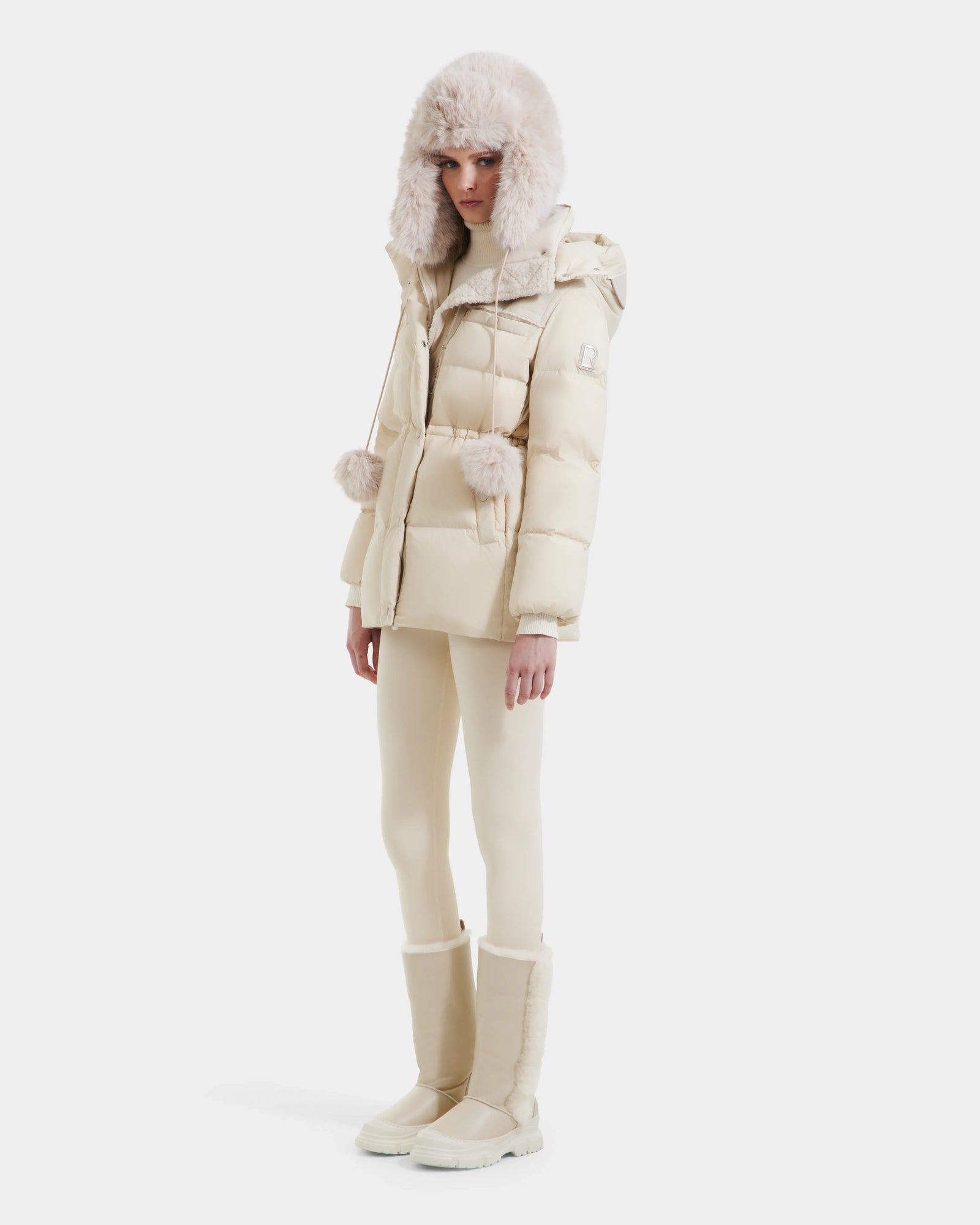 A woman stands against a white background wearing the Rudsak EMBER M-M - L125524 down puffer in Vanilla, featuring RDS-certified down, fur trim, a matching hat, slim pants, and tall beige boots.