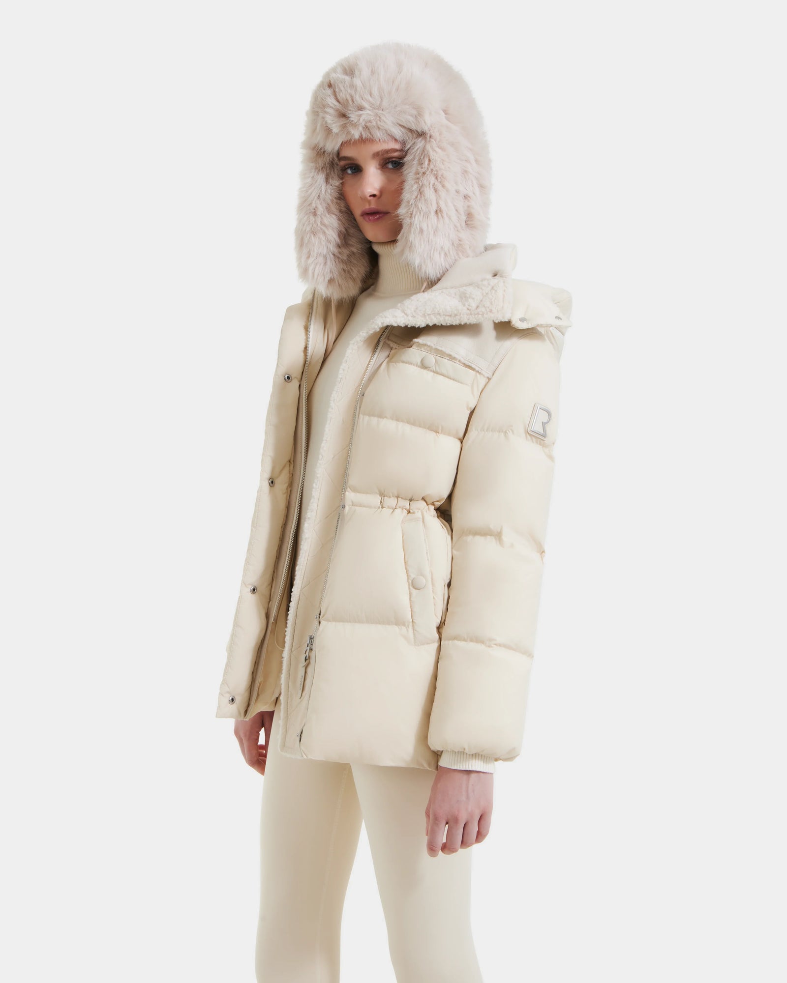 A woman wearing the Rudsak EMBER M-M - L125524 Vanilla down puffer jacket, a matching faux fur ear flap hat, and cream pants stands against a plain white background.