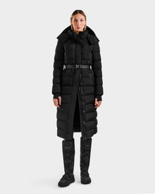 A woman faces forward against a plain light background, wearing the Rudsak FRIDA M-L - L125608 Black women’s long down puffer with a detachable hood, black pants, and matching black winter boots.