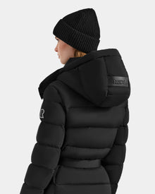 A woman is seen from behind wearing the Rudsak FRIDA M-M - L125607 Black quilted down puffer with a detachable hood and sleeve patch, plus a black beanie. The hood displays Rudsak branding, all set against a plain light background.