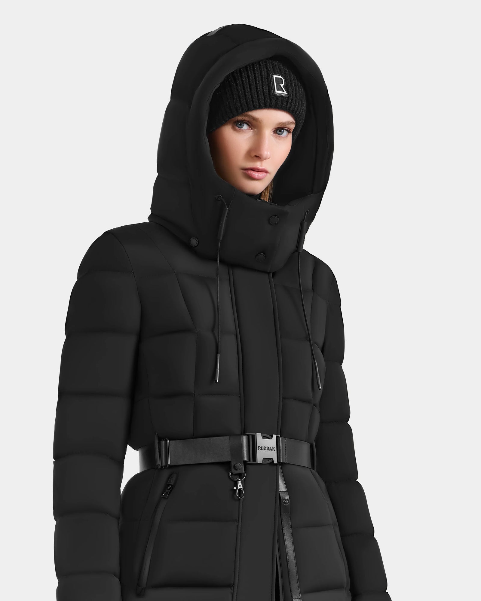 A person wears the Rudsak FRIDA M-M - L125607 Black women's quilted down puffer jacket with detachable hood and belt, paired with a black knit beanie, standing against a plain light background—a true winter essential.