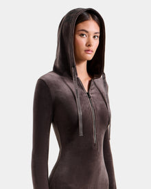 A woman with straight brown hair wears the Rudsak ISABELLA - L525502 Espresso, a fitted women’s velour hooded dress with a front zipper and long sleeves, looking forward neutrally against a plain light background.