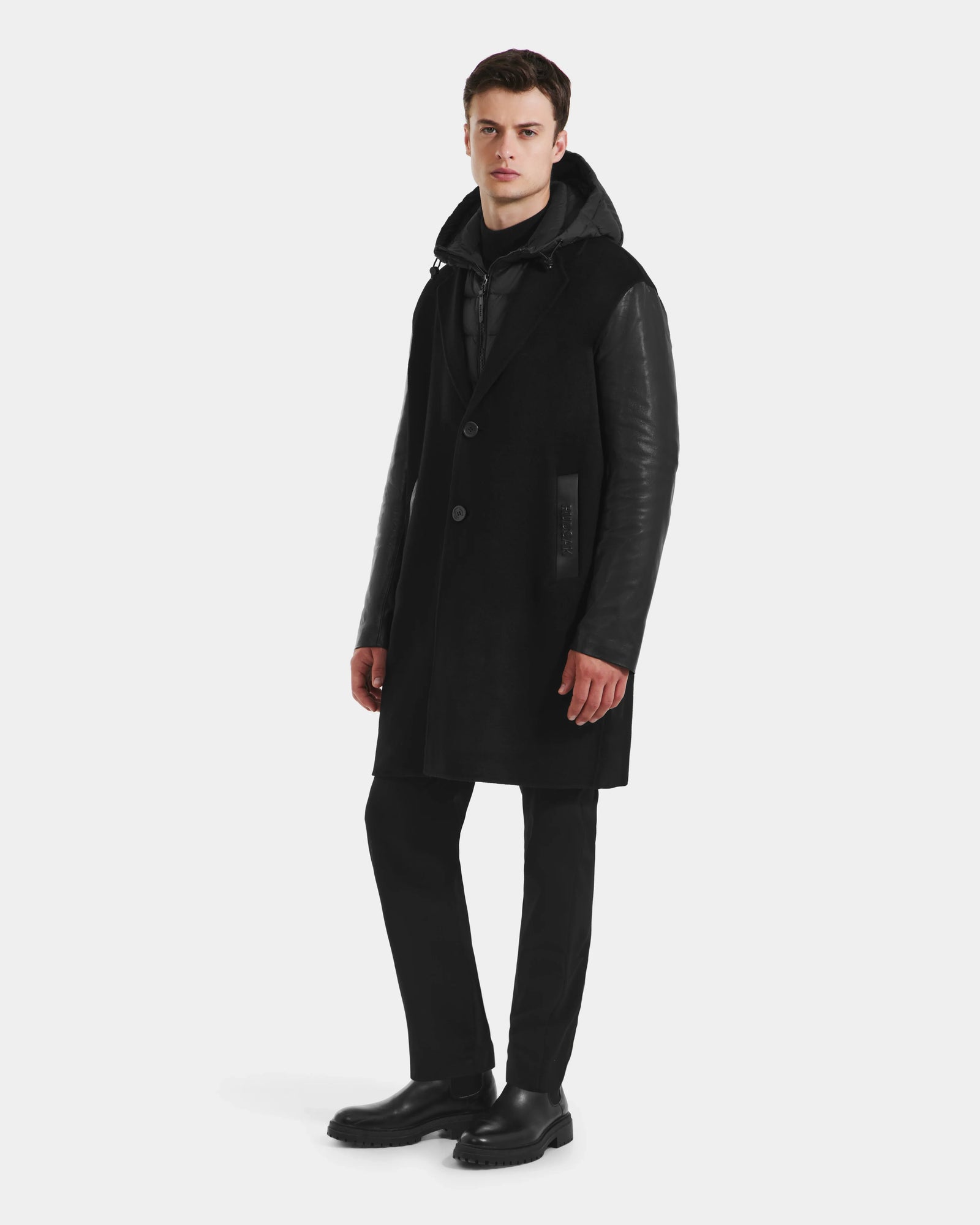 A man stands against a plain background wearing the Rudsak JESSE - M125504 Black men’s wool coat with leather sleeves, paired with black pants, black shoes, and a dark hoodie. He looks forward with a neutral expression.