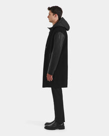 A man stands in profile against a white background, wearing the Rudsak JESSE - M125504 Black men’s wool coat with leather sleeves, styled with black pants, boots, and a down-insulated hood.