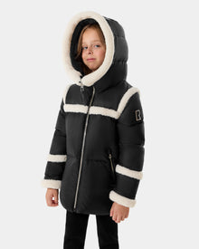 A young child models the Rudsak JULIANA K - K125550 Black puffer jacket with white faux fur trim on the hood, shoulders, chest, and cuffs, standing confidently against a plain light-gray background.