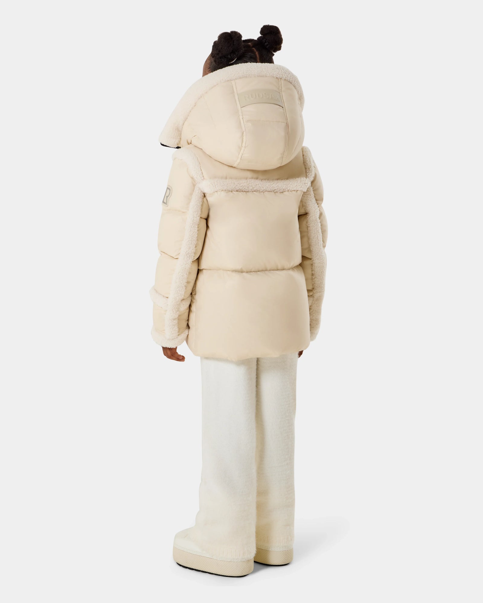 A child faces away, wearing the Rudsak JULIANA K - K125550 Vanilla light beige puffer jacket with hood, cream pants, and matching boots against a plain white background.