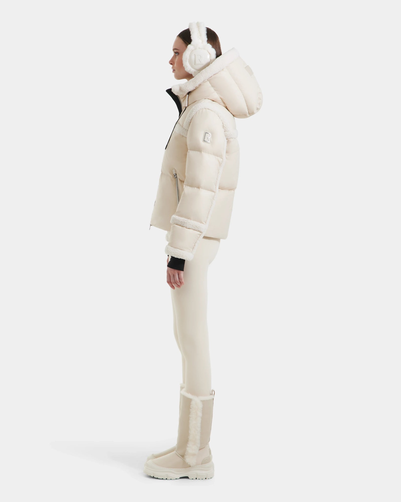 A woman in profile wears the Rudsak JULIANA - L124509 Vanilla short down puffer jacket with a hood, paired with matching leggings, earmuffs, and winter boots—an essential winter look—set against a plain white background.