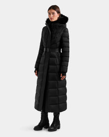 A woman models the Rudsak JULIETA M-L - L125559 Black long puffer coat with a black fur-trimmed hood and matching belt, paired with black lace-up boots, against a plain light gray background.