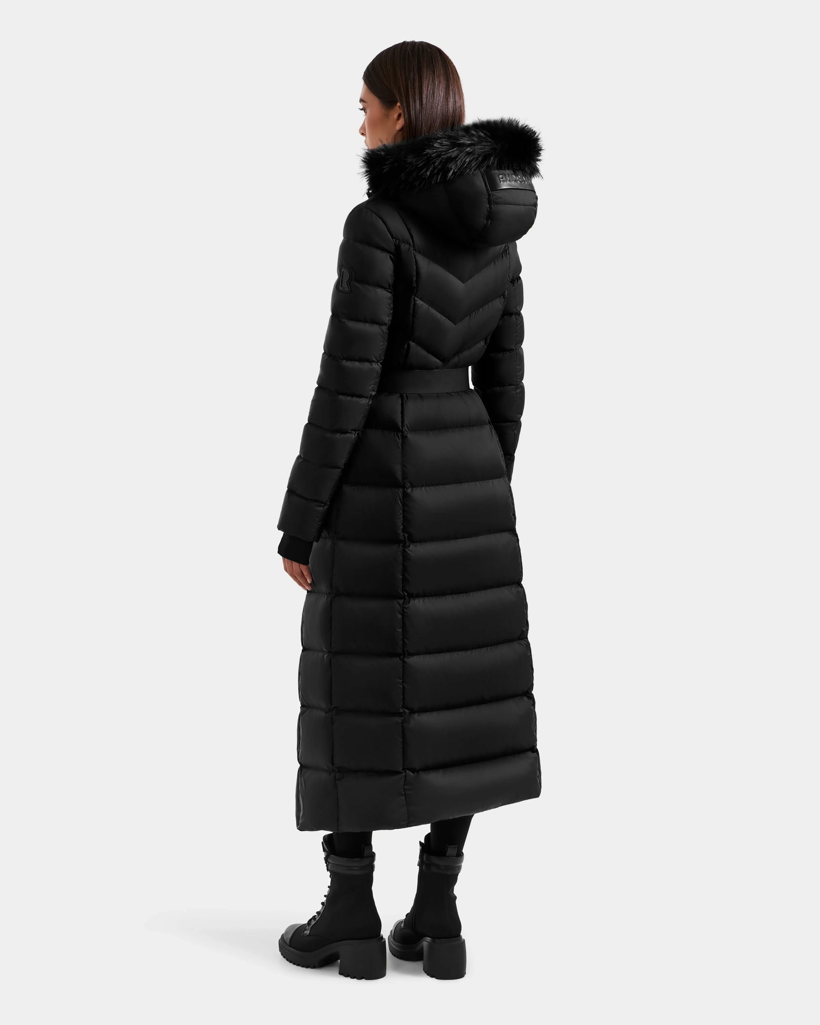 A woman faces away in the Rudsak JULIETA M-L - L125559 Black maxi down puffer featuring a weather-resistant shell, fur-trimmed hood, fitted waist, and horizontal stitching, paired with black lace-up boots. Plain light gray background.