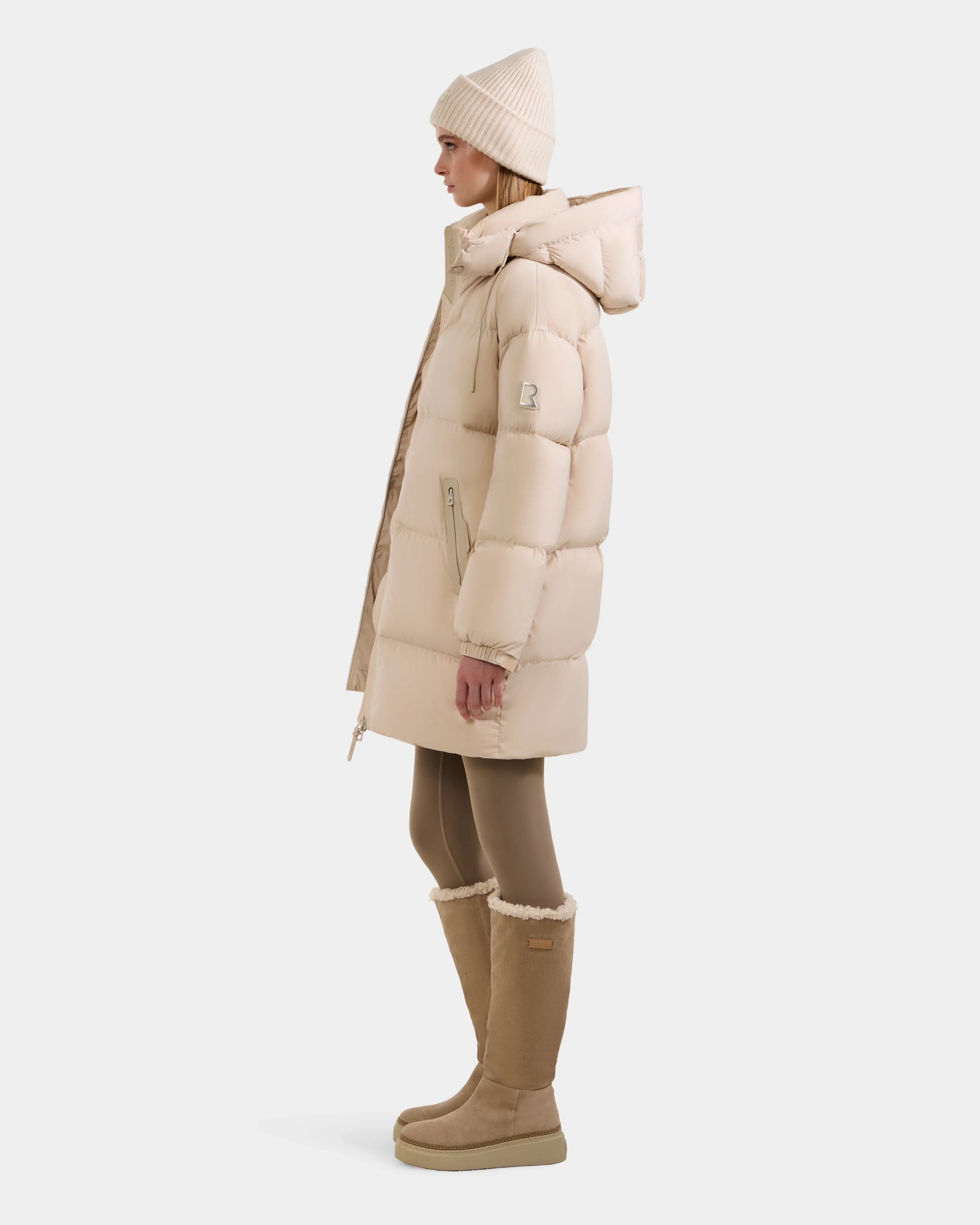 A person in profile wears the Rudsak KARLEE L - 8125682 Vanilla women’s long down puffer with vegan leather trims, a matching knit hat, tan leggings, and tan boots, posed against a plain white background.