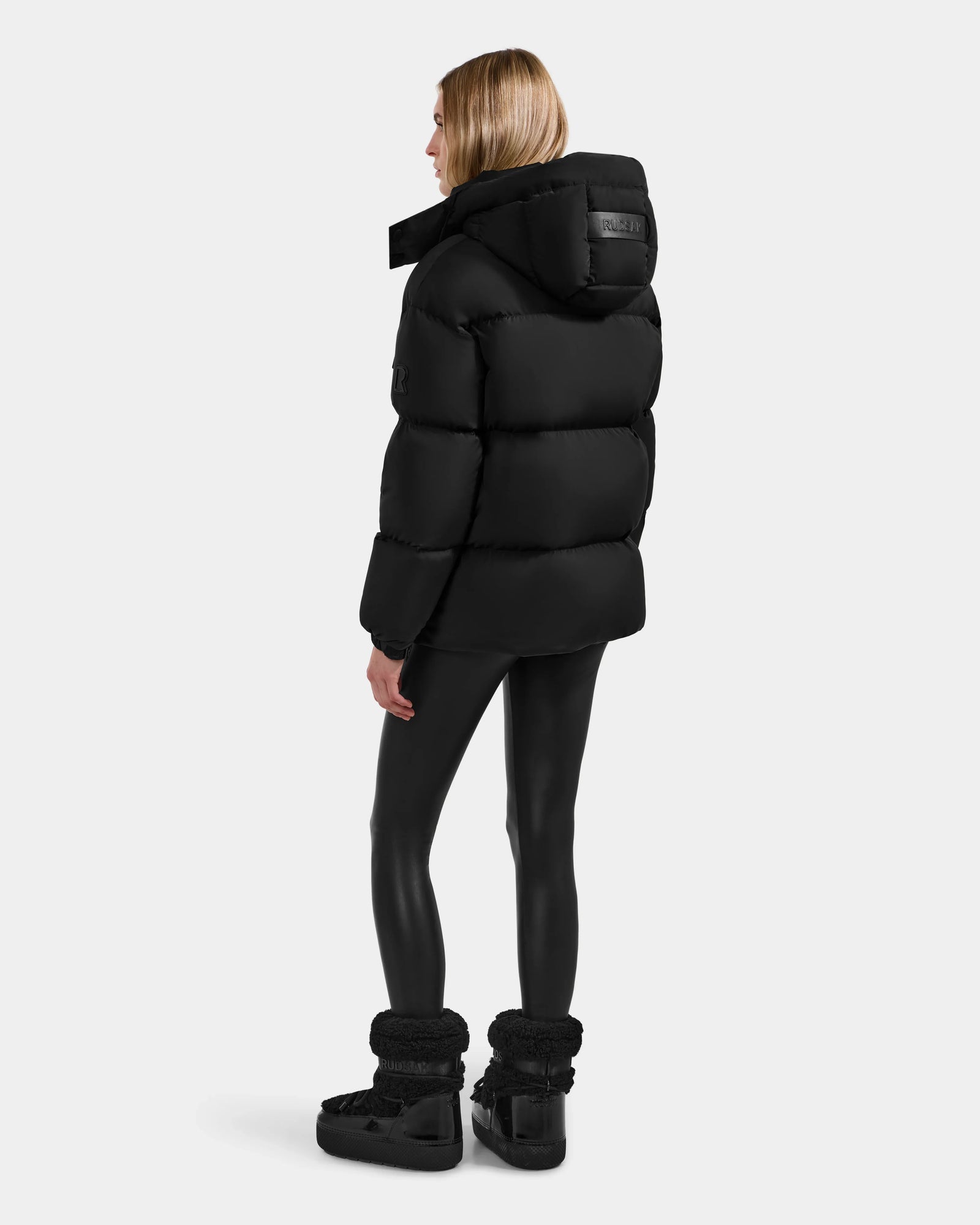 A woman with straight blonde hair faces away, wearing the Rudsak KARLEE M - 8125681 Black mid-length women’s down puffer jacket, black leggings, and black winter boots, exuding winter warmth against a plain white background.