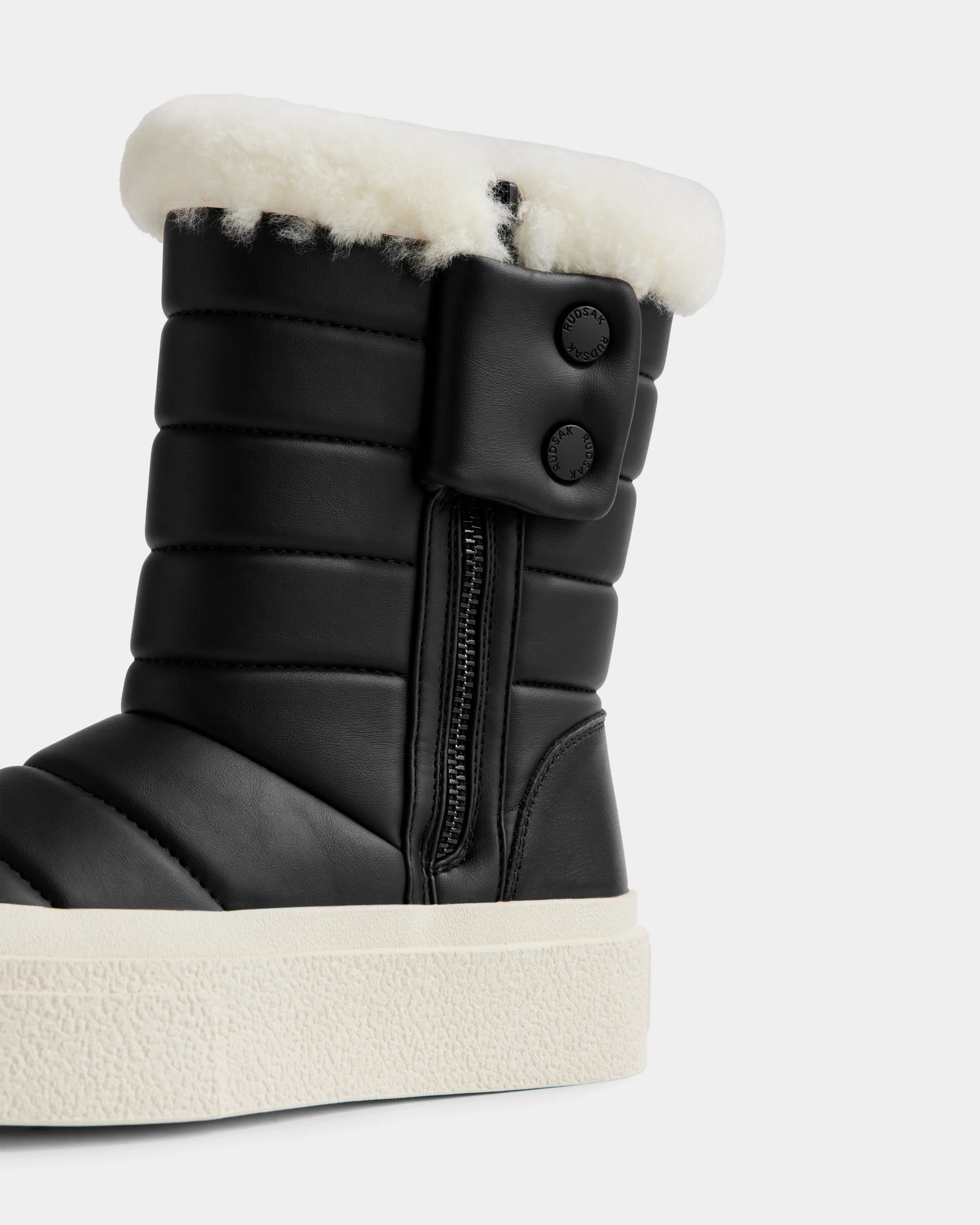 A close-up of Rudsak’s KATHIE - L225516 Black women’s mid boot, featuring black quilting, a side zipper, two buttons, white faux fur lining, and a thick white textured rubber sole.