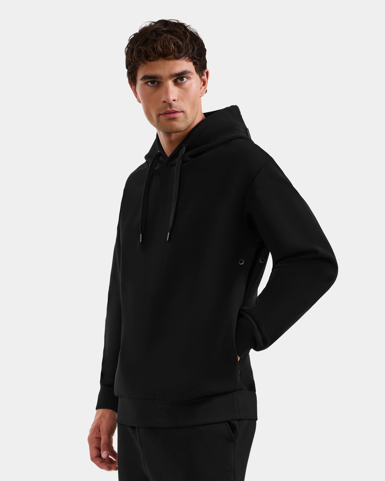 A young man with short curly hair models the Rudsak LEROY - U525509 Black M hoodie with drawstrings and black pants, standing against a light gray background, one hand in his pocket, gazing slightly to the side.