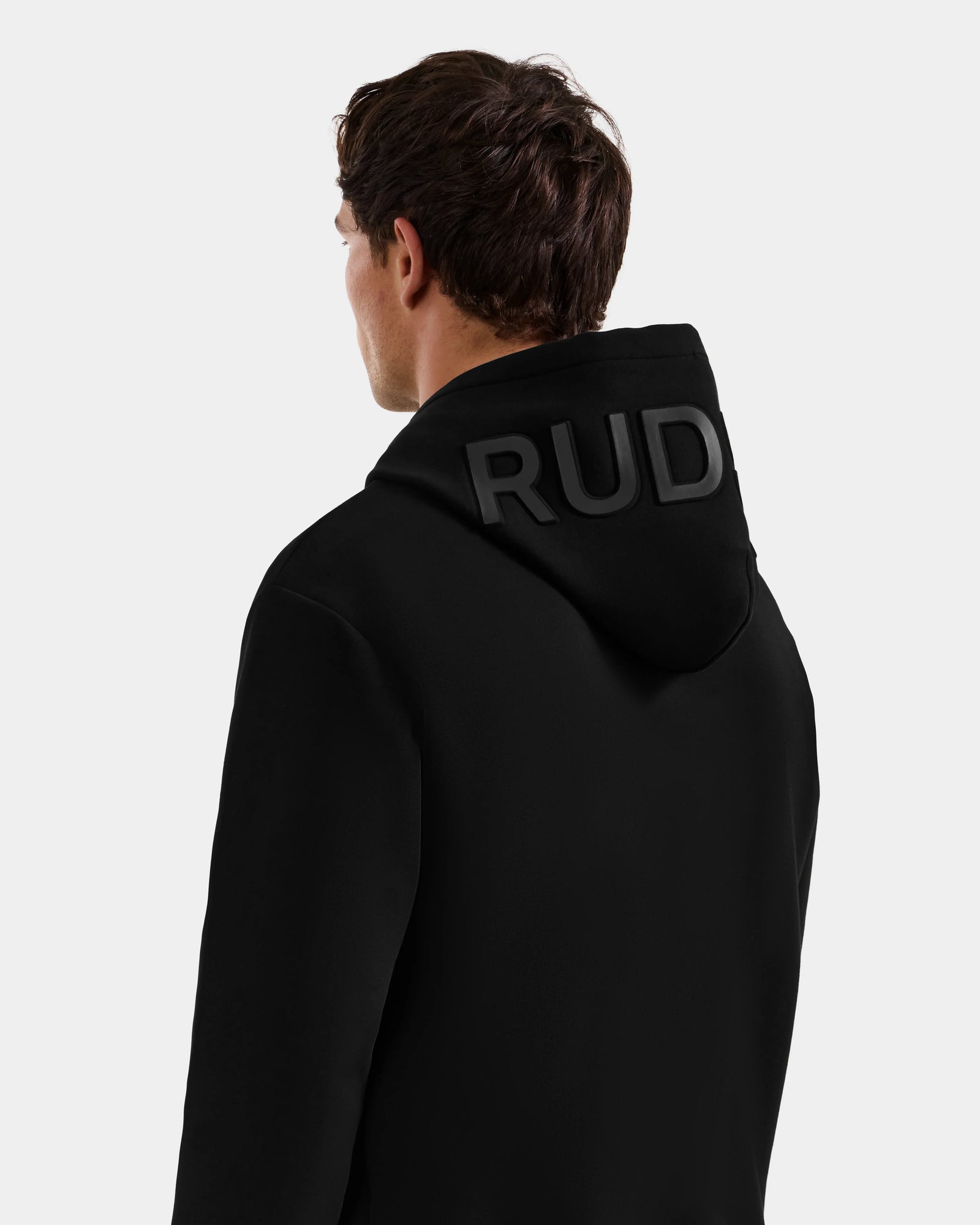A person with short brown hair faces away, wearing the Rudsak LEROY - U525509 Black M unisex hoodie, featuring "RUD" embossed in large letters on the hood, set against a plain light background.