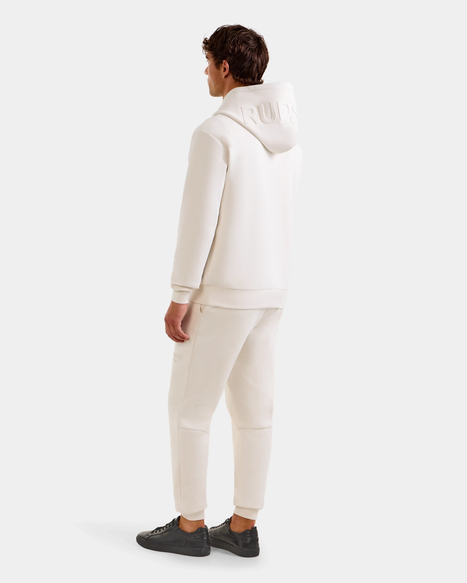 A person faces away, wearing the Rudsak LEROY - U525509 Vanilla M unisex cotton-blend hoodie tracksuit with embossed hood text and black sneakers, set against a plain white background.