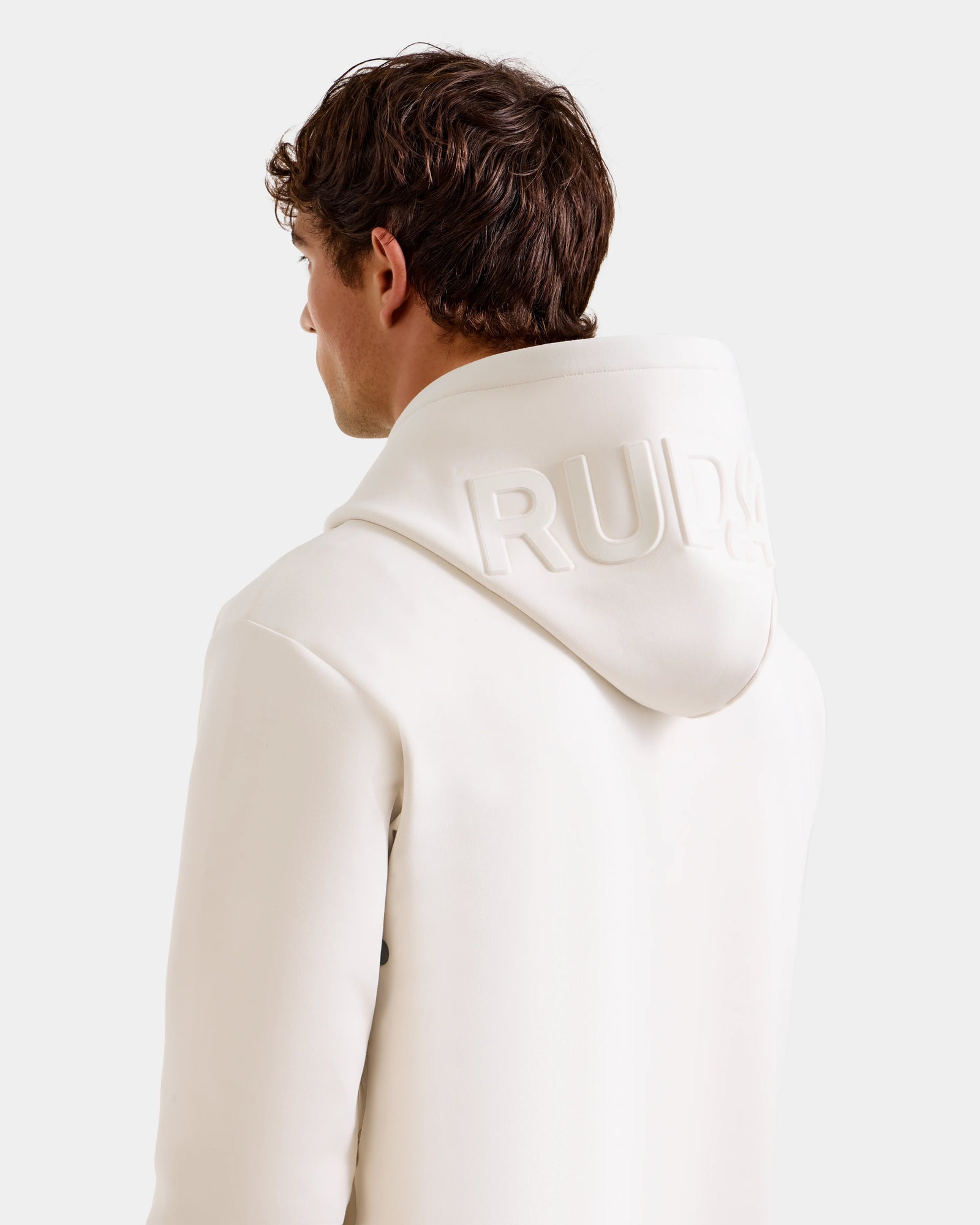 A person with short brown hair, seen from behind, wears the Rudsak LEROY - U525509 Vanilla M hoodie with the hood up, featuring embossed "RUDA" text. The background is plain and light-colored.