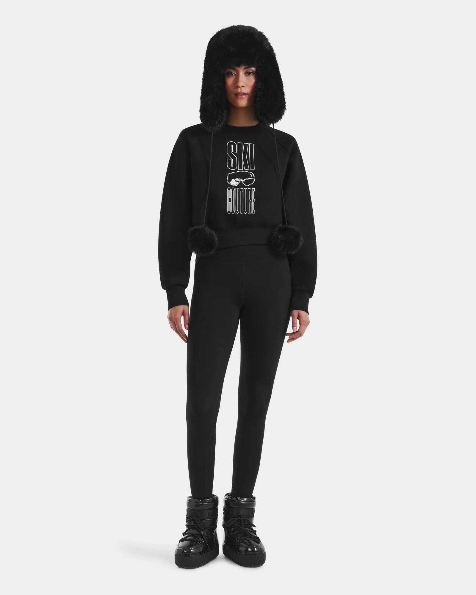 A person wears Rudsak's LIDDIE - L525505 Black: an all-black après-ski outfit featuring a furry trapper hat, SKI COUTURE women’s sweatshirt, fitted leggings, and puffy snow boots, standing against a white background.