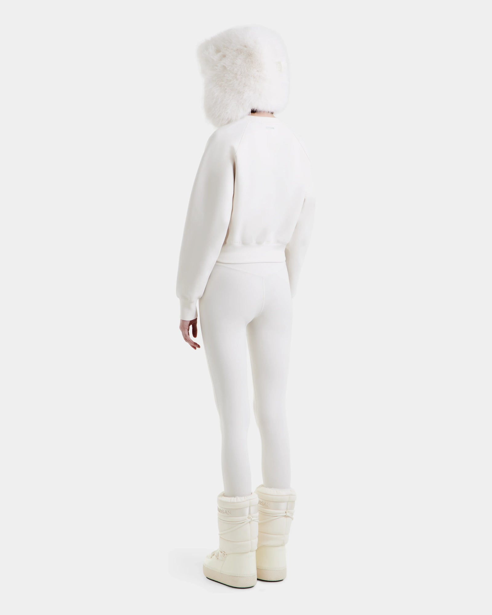 A person stands facing away, dressed in the Rudsak LIDDIE - L525505 Vanilla all-white winter outfit, featuring a furry hat, fitted jacket, leggings, and puffy boots against a plain light background.