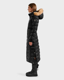 A woman in profile wears the Rudsak LILITH L - L125528 Black puffer coat with fur-trimmed hood, quilted design, and side snaps, paired with black lace-up boots against a plain light background.