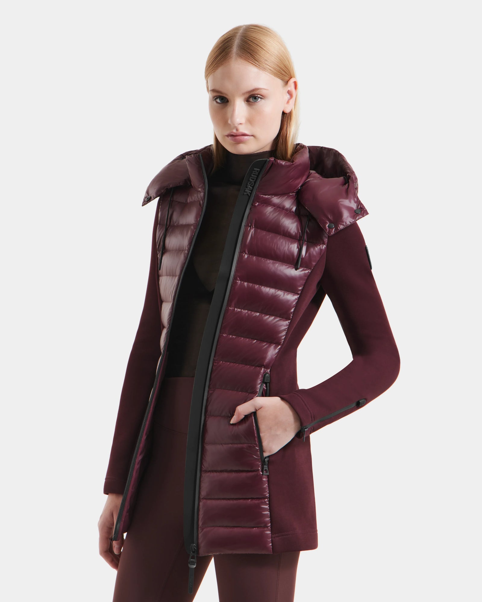 A woman with straight blonde hair models the Rudsak MARRY S - L125521 Wine, a weather-resistant burgundy coat with quilted, shiny panels and a high collar. The glossy jacket is partially unzipped over a dark top, set against a plain white backdrop.