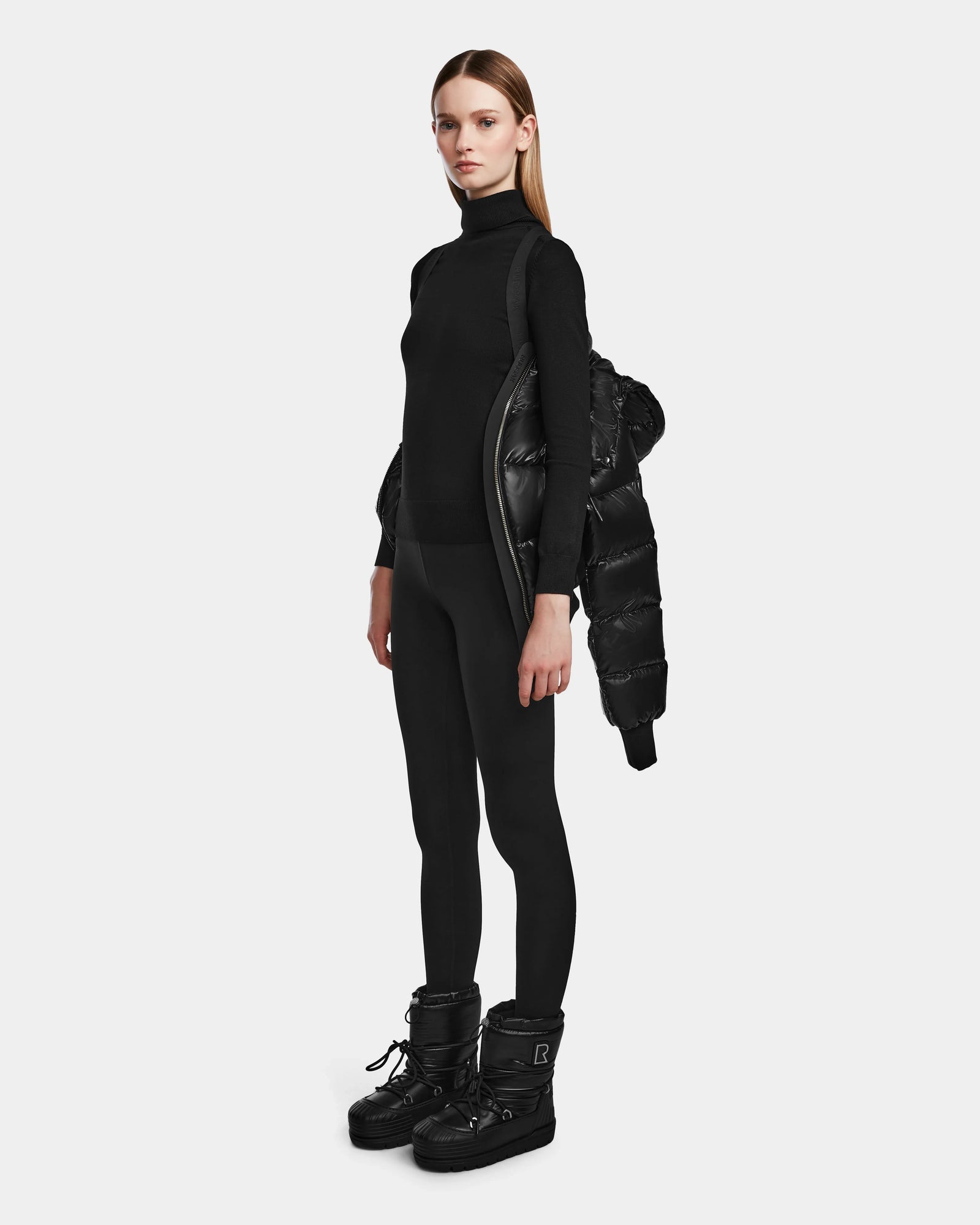 A woman stands against a plain background in a black turtleneck, leggings, and boots, with the Rudsak MAUDE Q-S - L124553 Black RDS-certified down jacket draped over one shoulder.