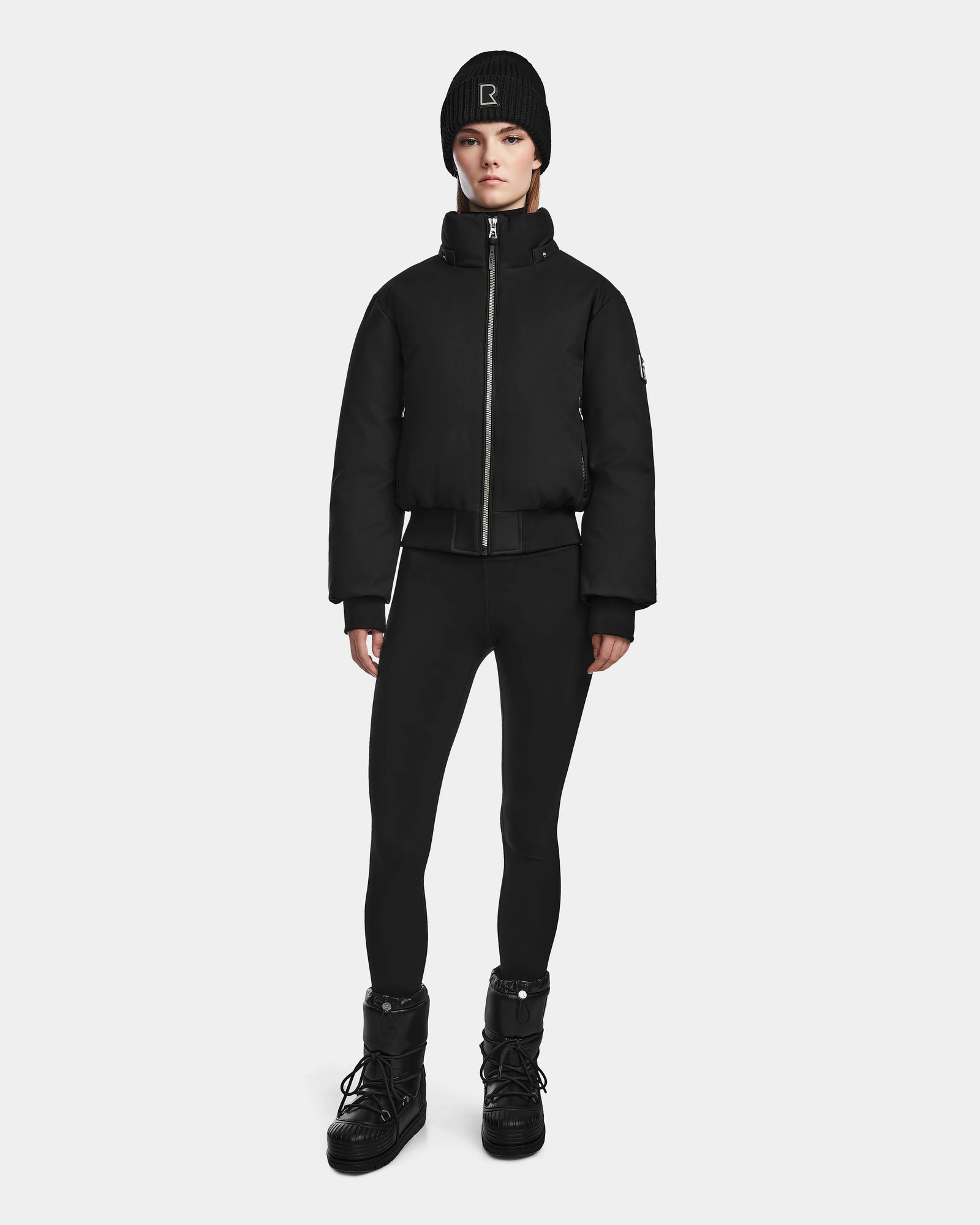 A person stands facing forward, wearing the Rudsak MAUDE - L124522 Black/Black women's down bomber, paired with black leggings and black boots against a plain light background.