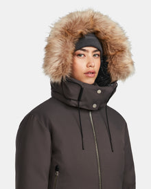A woman models the Rudsak MAUDE - L124522 Grey down bomber, zipped up with silver snaps and drawstrings. The classic winter jacket features a fur-lined hood, premium down insulation, and is paired with a gray beanie.