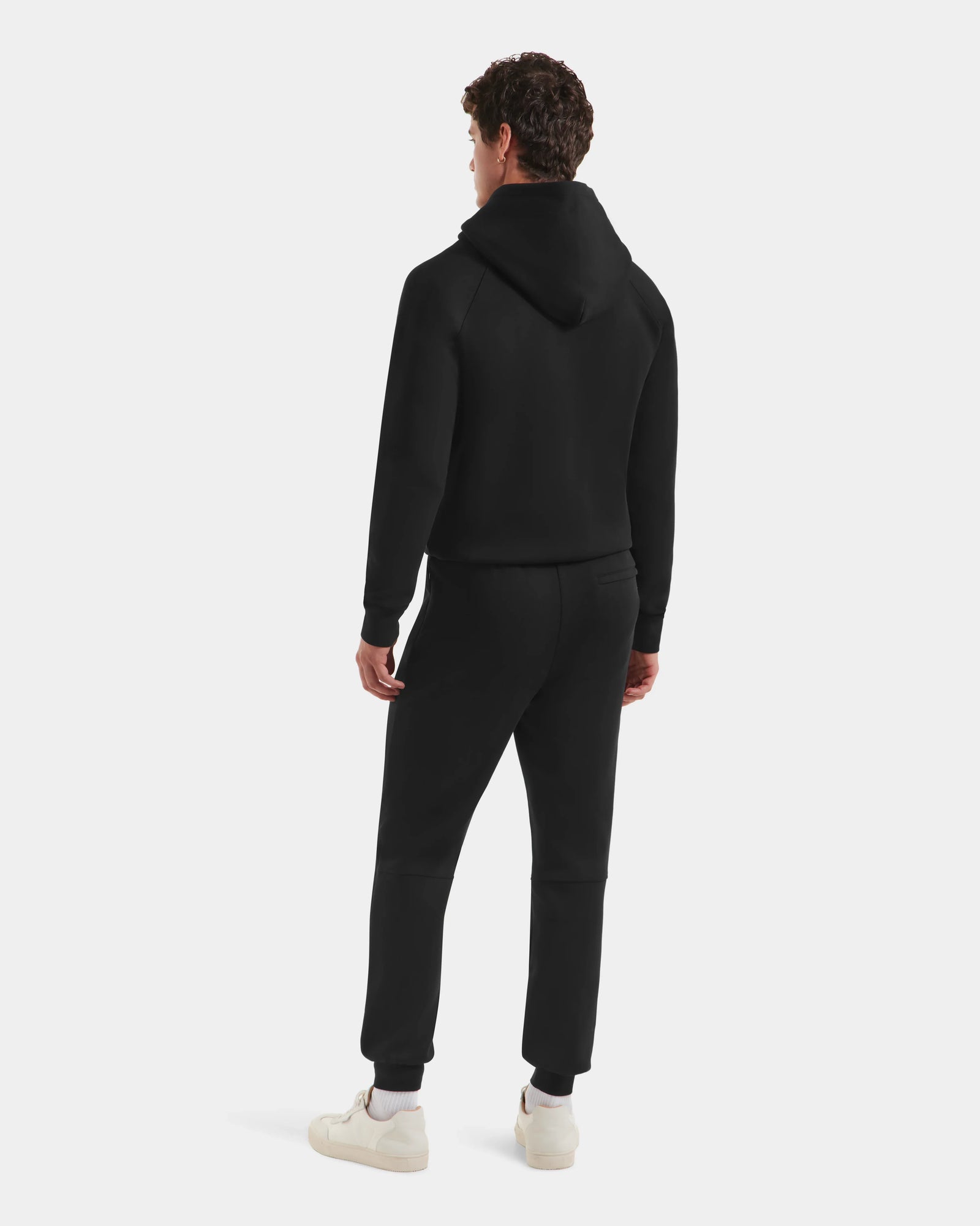A person faces away, wearing the Rudsak MAXTON - U525007 Black M hoodie and unisex jogger pants with white sneakers, set against a plain light background.