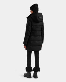 A person wearing Rudsak’s MIRIAM MW - L125604 Black RDS-certified down puffer coat with a hood, a black knit hat, black leggings, and fur-trimmed black boots stands facing away against a plain light gray background.