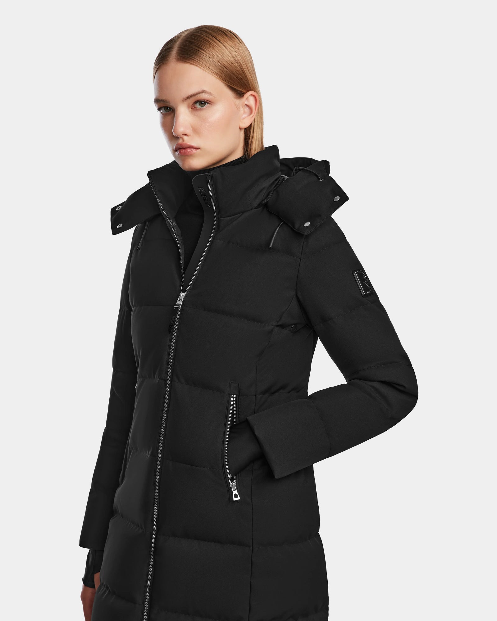 A woman with straight blonde hair models the Rudsak MIRIAM - L123544 Black, a long women’s down puffer winter coat with a high collar and zippered pockets, standing against a plain light background—a winter wardrobe staple.
