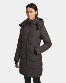 A woman models the Rudsak MIRIAM - L123544 Grey long down puffer coat with hood, weather-resistant shell, front zipper, zippered pockets, and upper sleeve patch against a white background.