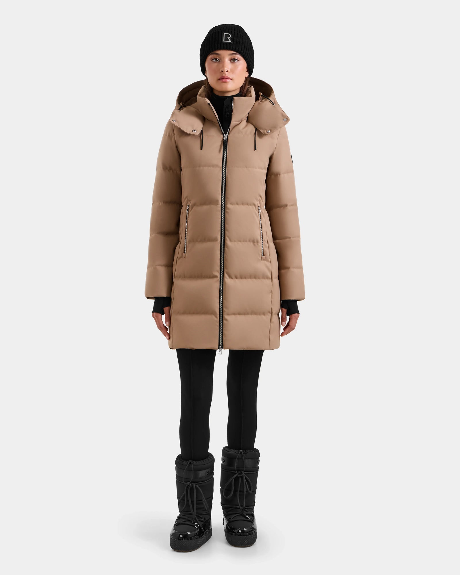 A woman faces forward in a plain white setting, wearing the Rudsak MIRIAM - L123544 Sand women’s down puffer winter jacket with a hood, paired with black leggings, black winter boots, and a black beanie.