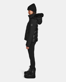 A person stands in profile wearing the Rudsak MIRRY M - L124533 Black RDS-certified down coat with a fur-lined hood, black leggings, a black beanie, and black boots against a plain white background.