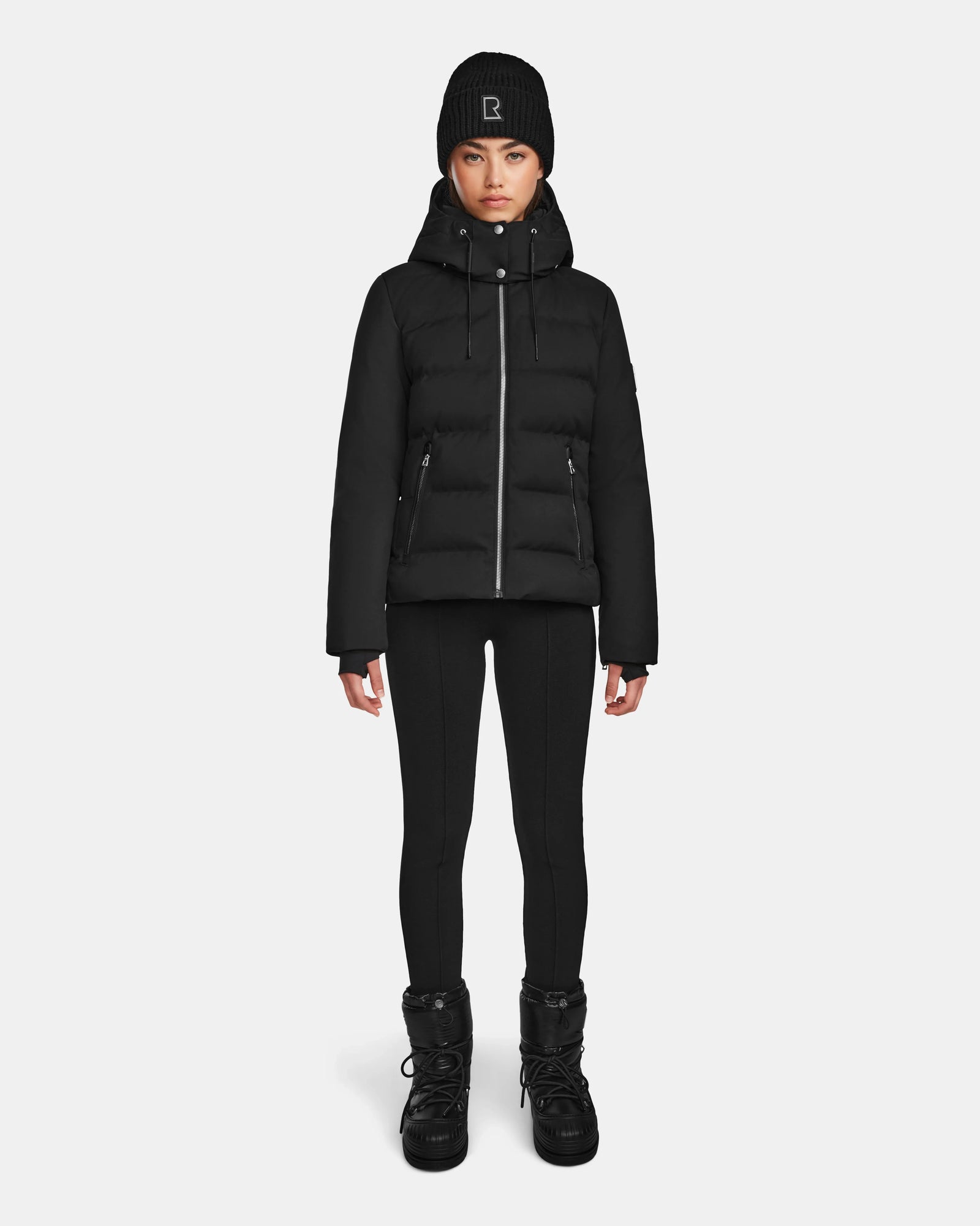 A person stands facing forward against a plain white background, wearing the Rudsak MIRRY M - L124533 Black puffer jacket, black leggings, black winter boots, and a black knit beanie with a small white logo.