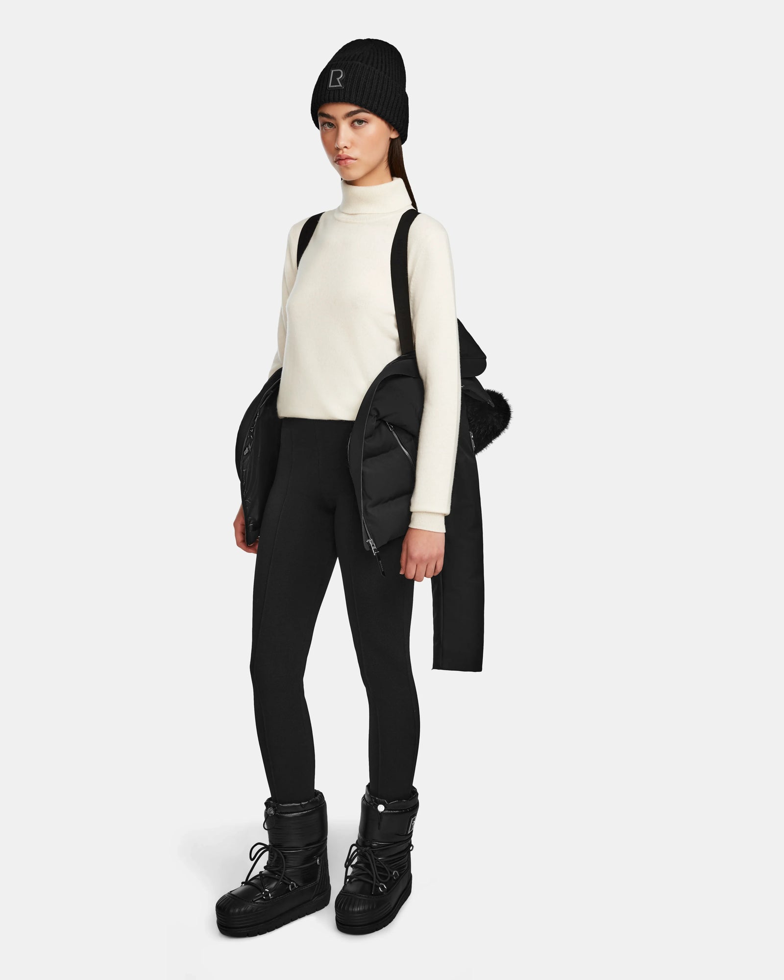 A woman stands facing the camera against a white background, carrying the Rudsak MIRRY M - L124533 Black women's quilted down coat over her shoulders. She wears a black beanie, white turtleneck sweater, black leggings, and black winter boots.