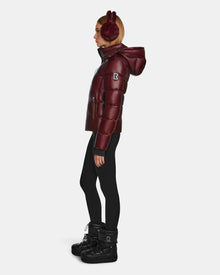 A woman in profile wears burgundy earmuffs, black leggings, black winter boots, and a Rudsak MIRRY S - L123545 Wine quilted down coat with hood, set against a plain white background.