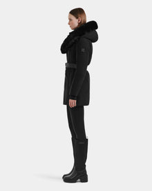 A woman stands in profile against a light background, wearing the Rudsak MODA M - L125555 Black winter coat with a large faux fur-trimmed hood, black leggings, knee-high boots, and a vegan leather belt at the waist.
