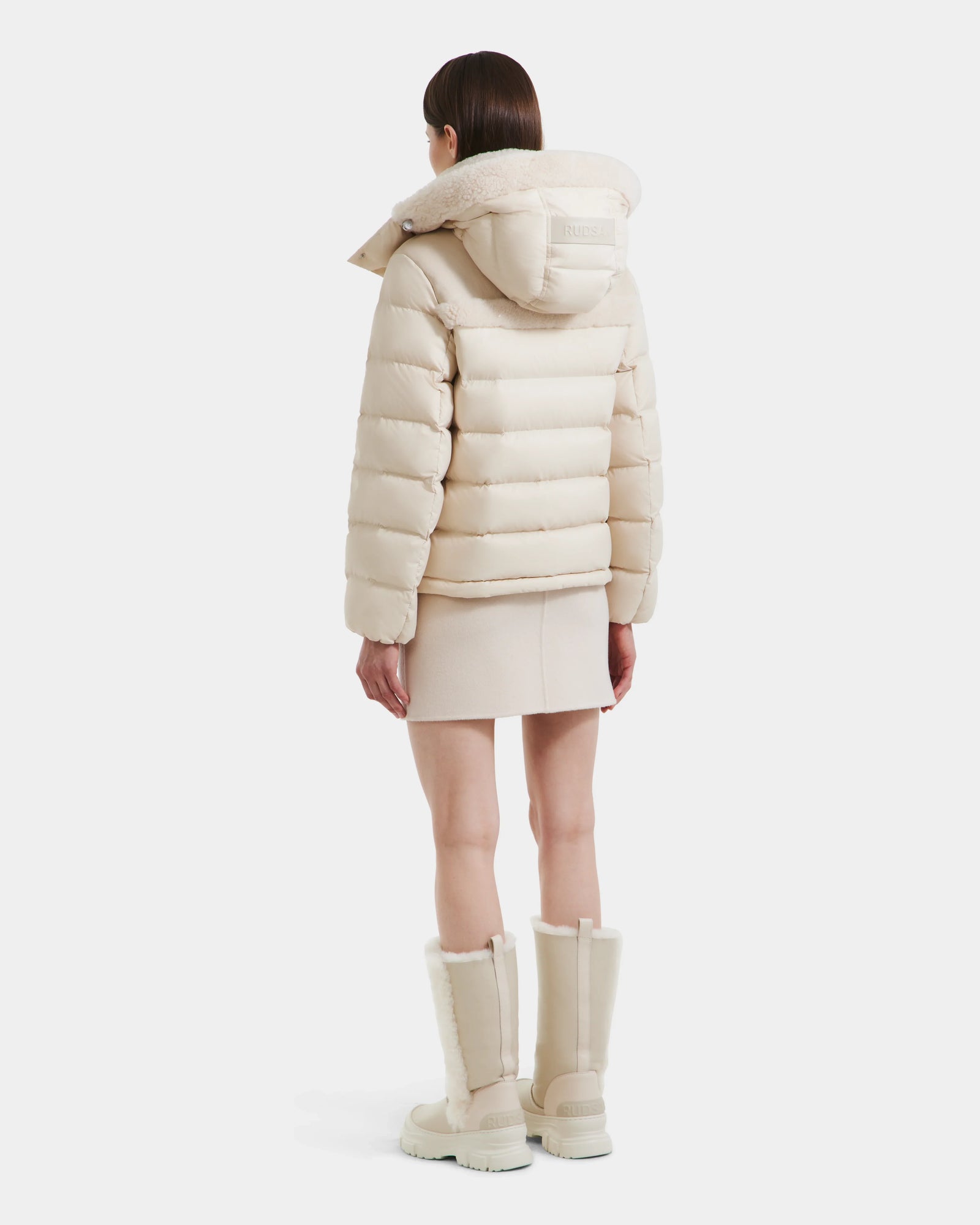 A woman faces away, modeling the Rudsak SANDRA - 8125612 Vanilla women's shearling puffer jacket with lightweight insulation, styled with a matching short skirt and fur-trimmed knee-high boots in light beige against a white background.