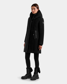 A person stands against a plain background wearing the Rudsak SARINA - L125550 Black women’s down parka with a large collar, paired with black leggings and black fur-trimmed lace-up boots. Their hands are at their sides and they have a neutral expression.