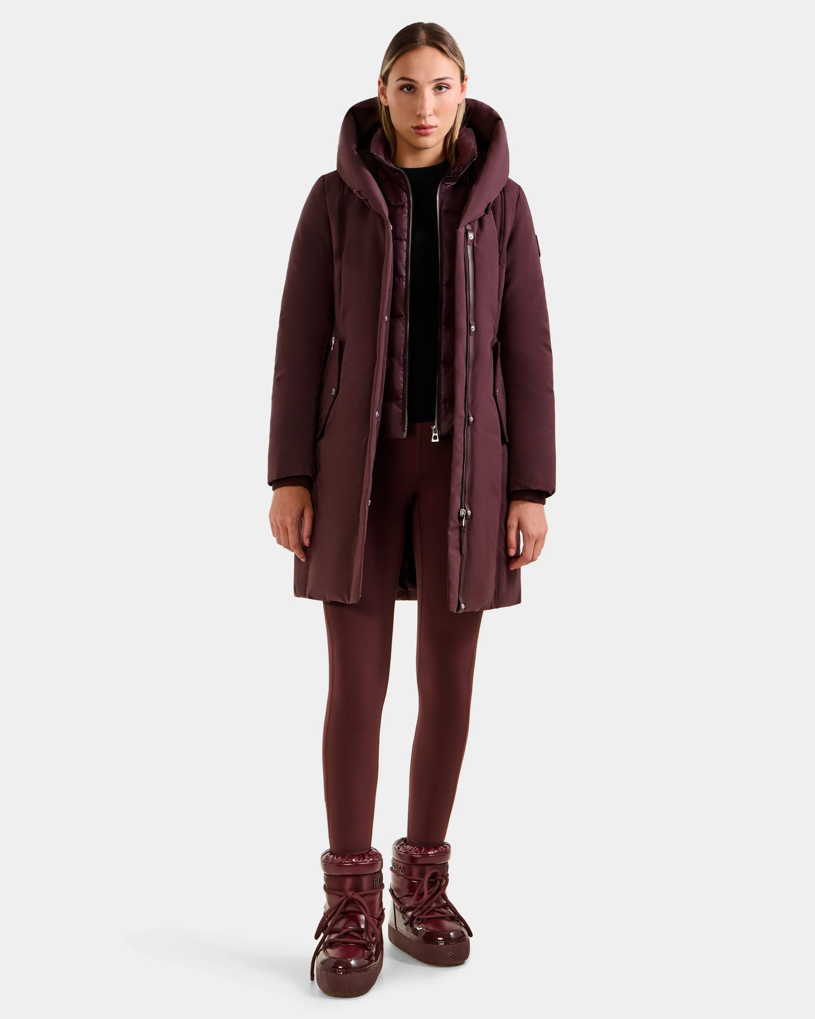 A woman faces forward wearing the Rudsak SARINA - L125550 Bordeaux down parka with matching leggings and shiny winter boots. Her hair is pulled back, her expression is neutral, and she stands against a plain white background.